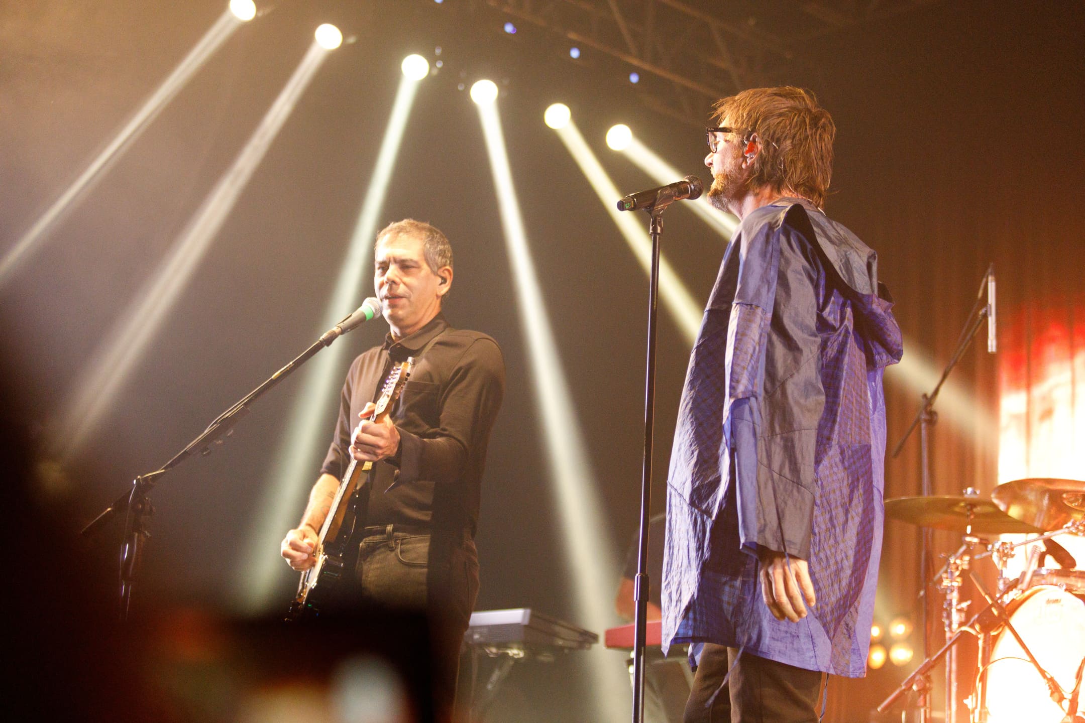 Dado Villa-Lobos e vocalista juntos no palco com iluminação de spots durante show As V Estações