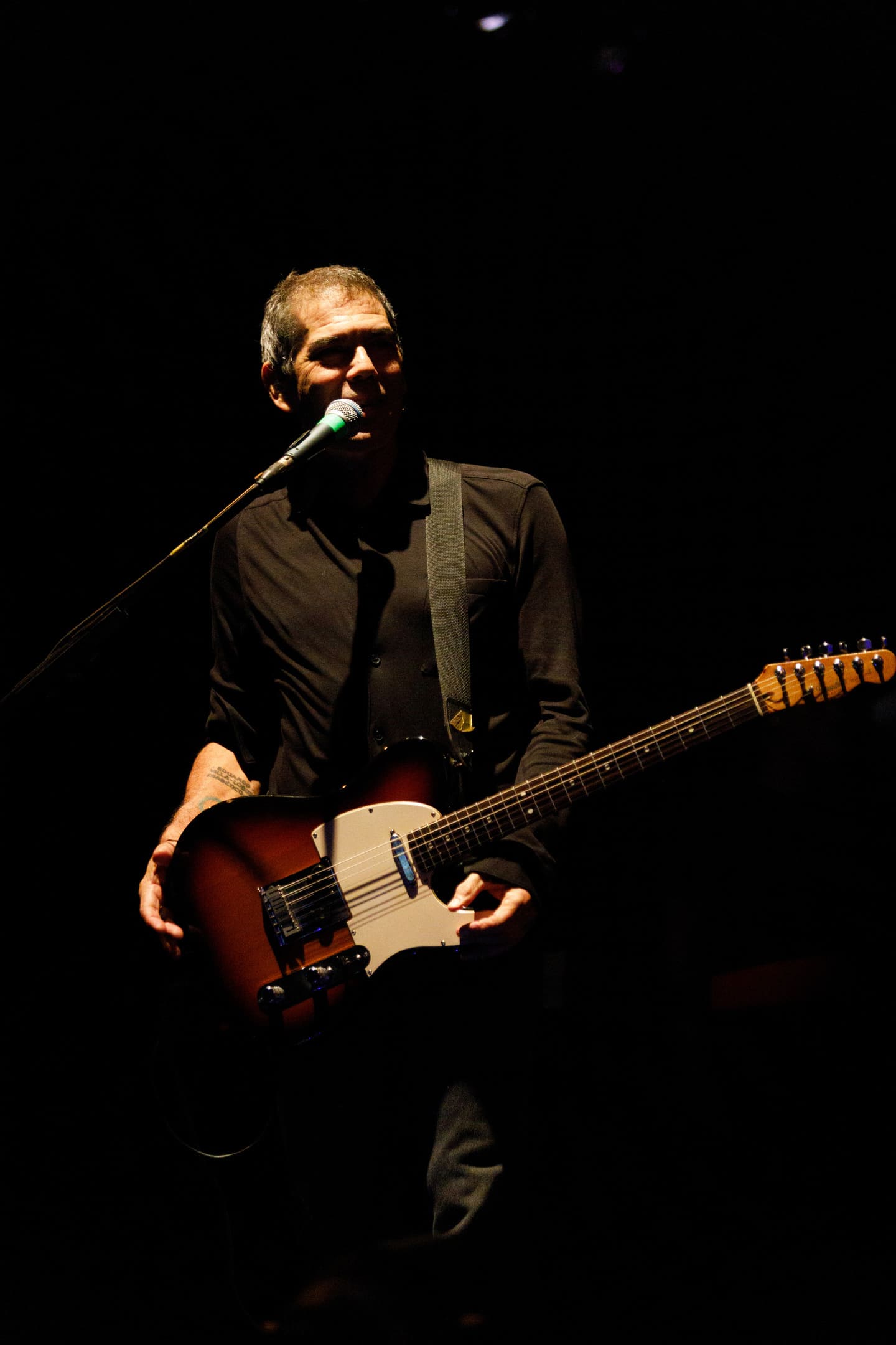 Dado Villa-Lobos com guitarra e microfone em iluminação dramática no show As V Estações Maringá